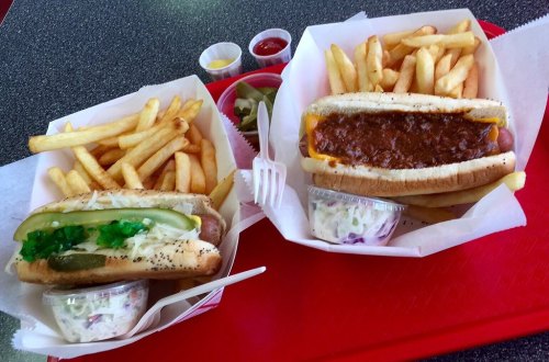 Two trays of food: one with a hot dog topped with chili and cheese, the other with a pickle-topped hot dog and fries.