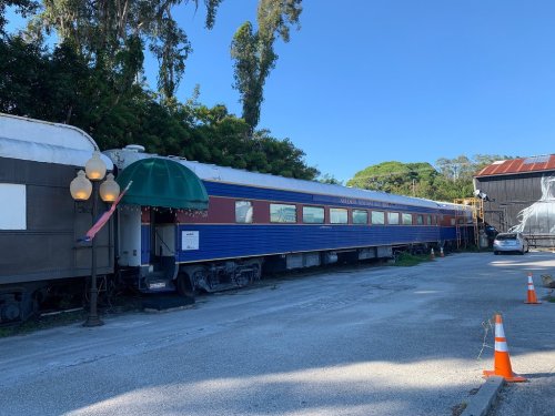 A vintage train car parked beside a road, surrounded by trees and a clear blue sky.