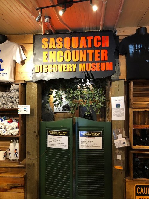 Sign for the Sasquatch Encounter Discovery Museum, featuring a rustic design and green foliage.