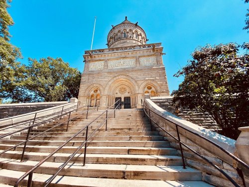 A grand stone building with a dome, featuring intricate carvings, surrounded by trees and a clear blue sky.