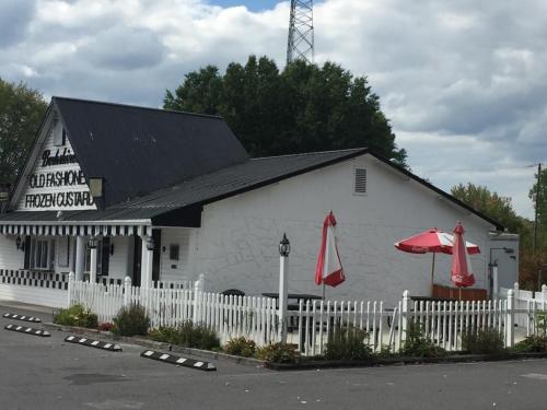 A quaint frozen custard shop with a white exterior, red umbrellas, and a picket fence, set against a cloudy sky.