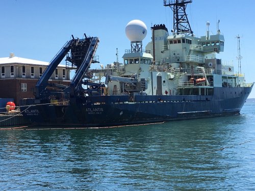 Research vessel Atlantis docked in calm waters, featuring a large crane and satellite equipment on its deck.