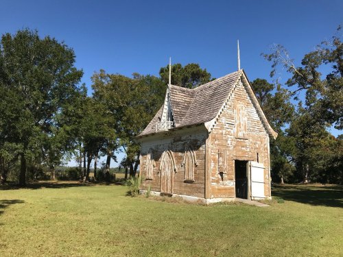 A small, weathered chapel with gothic features, surrounded by trees and a clear blue sky.