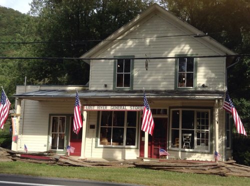 A general store with American flags, featuring a porch and wooden steps, set against a backdrop of trees.