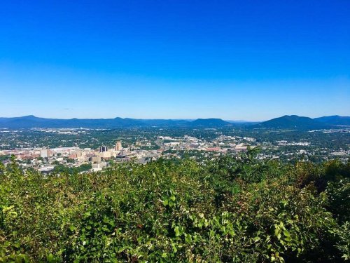 A panoramic view of a city surrounded by green hills under a clear blue sky.