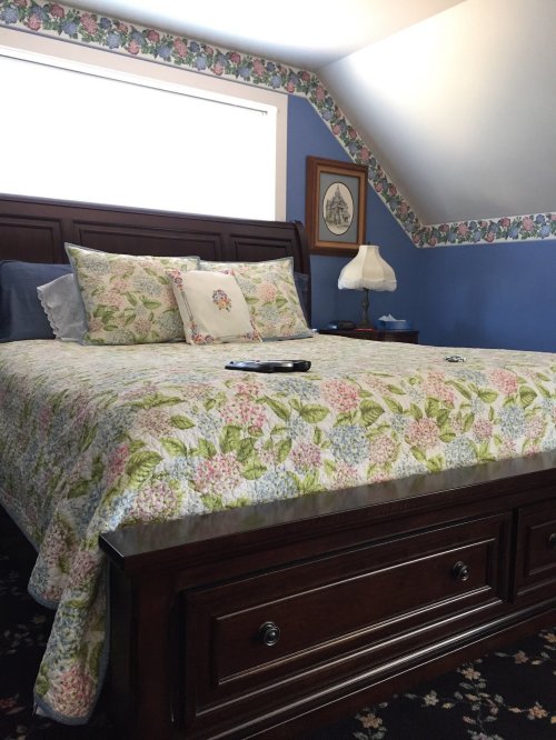 A cozy bedroom featuring a floral quilt, wooden bed frame, and blue walls with floral trim. A lamp sits on a nightstand.