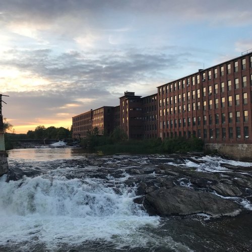 A river flows over rocks in the foreground, with a brick building and sunset in the background.