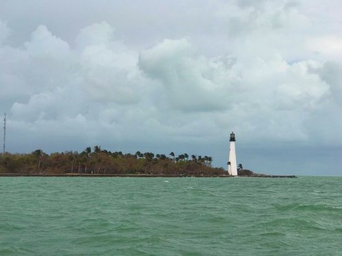 A lighthouse stands on a small island surrounded by green water and cloudy skies.