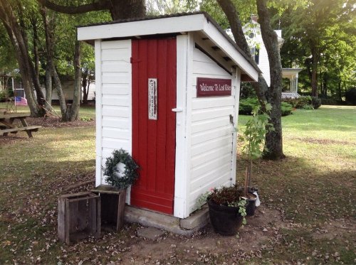 A small white shed with a red door, surrounded by greenery and potted plants, in a peaceful outdoor setting.