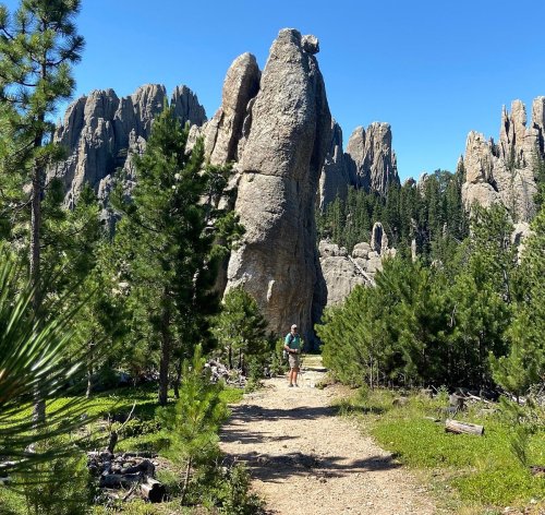 A hiker walks along a trail surrounded by tall rock formations and lush green trees under a clear blue sky.