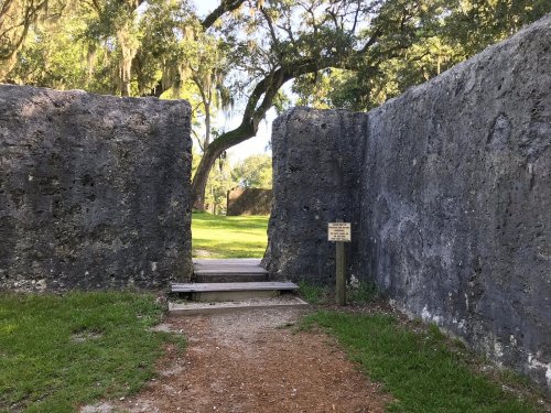 A pathway leads through weathered stone walls, surrounded by greenery and trees, with a sign nearby.