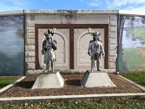 Two metallic statues of Indigenous figures stand in front of a mural depicting a natural landscape.