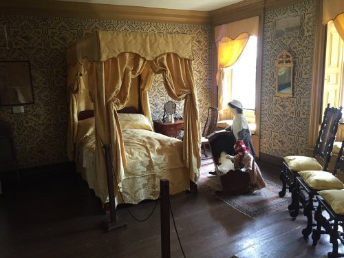 A vintage bedroom with a canopy bed, ornate wallpaper, and a woman in period attire sitting by a window.