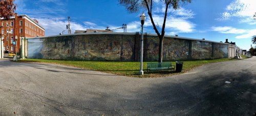 A panoramic mural on a building wall, depicting a vibrant landscape with trees and figures, surrounded by a park setting.