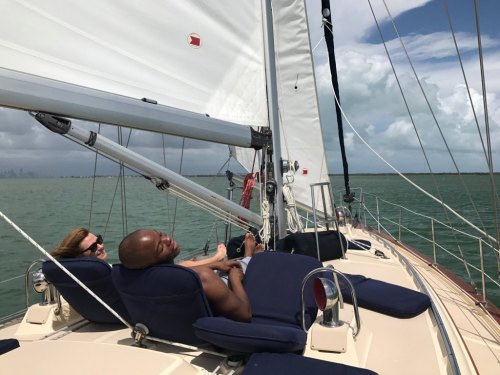 Two people relax on a sailboat, enjoying the view of the water and clouds on a sunny day.