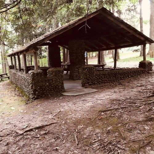 A rustic stone pavilion surrounded by trees, with picnic tables and a natural, wooded setting.