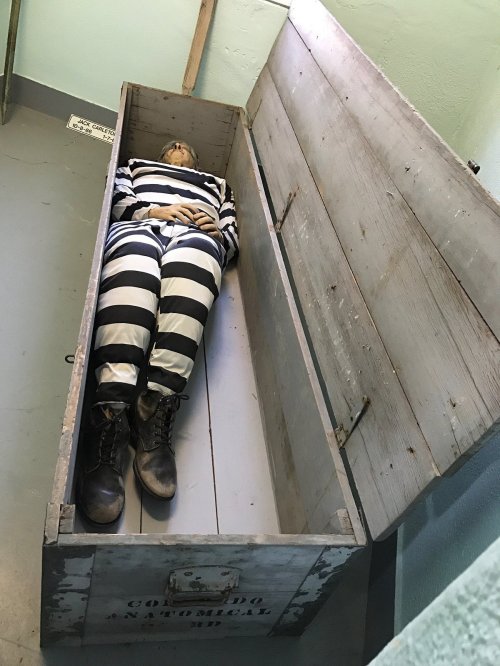 A lifelike mannequin in a striped prison outfit lies inside an open wooden coffin.