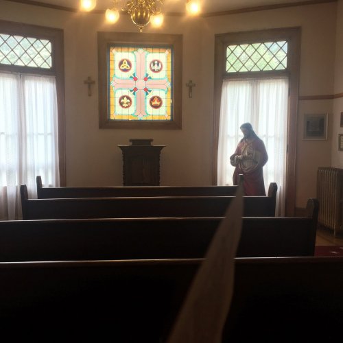 Interior of a small chapel with wooden pews, a stained glass window, and a statue of a figure near the altar.