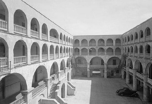 Black and white image of a courtyard with arched balconies and a vintage car parked below.