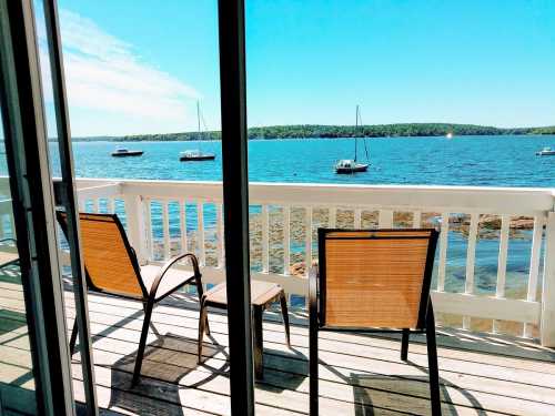 Two chairs on a deck overlooking a calm lake with sailboats and green trees in the background under a clear blue sky.