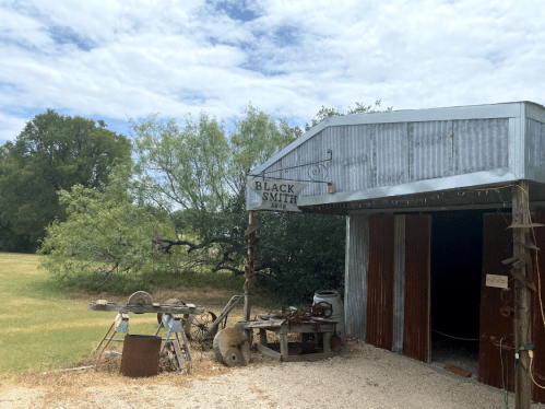A rustic blacksmith shop with a sign, surrounded by greenery and tools outside.