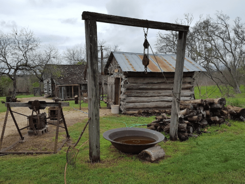 A rustic scene featuring a log cabin, a well with a pulley, and a stack of firewood in a grassy area.