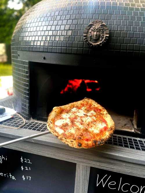 A freshly baked pizza on a peel, emerging from a wood-fired oven with a welcoming sign nearby.
