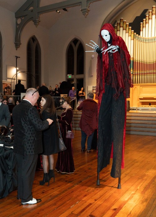 A tall, eerie figure in a red cloak and mask stands among elegantly dressed guests in a grand hall.