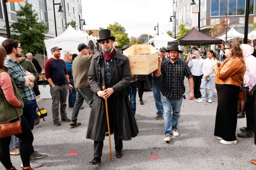 A man in a top hat and coat walks with a cane, while another carries a wooden box at a lively outdoor event.