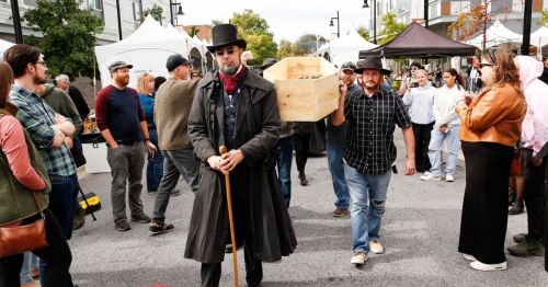 A crowd at an outdoor event, with a man in a top hat and coat walking ahead, carrying a wooden box.