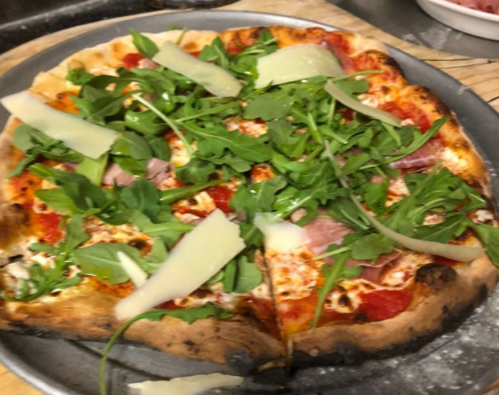 A freshly baked pizza topped with tomato sauce, arugula, prosciutto, and shaved cheese on a metal tray.