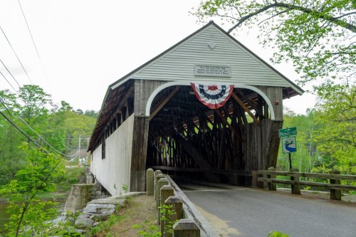 A historic covered bridge with wooden beams, decorated with bunting, surrounded by greenery and a road sign nearby.