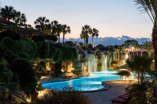 A serene pool area with waterfalls, surrounded by lush greenery and palm trees, illuminated at dusk.