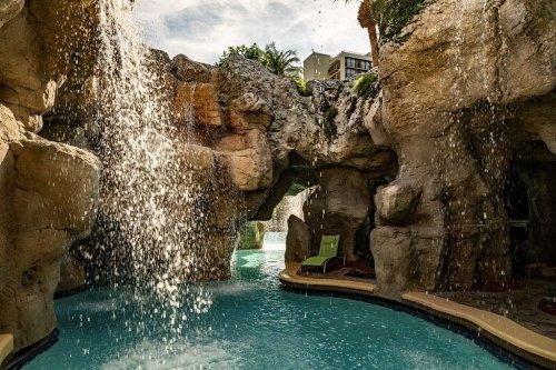 A serene pool area with a waterfall cascading over rocky formations, surrounded by lush greenery and lounge chairs.