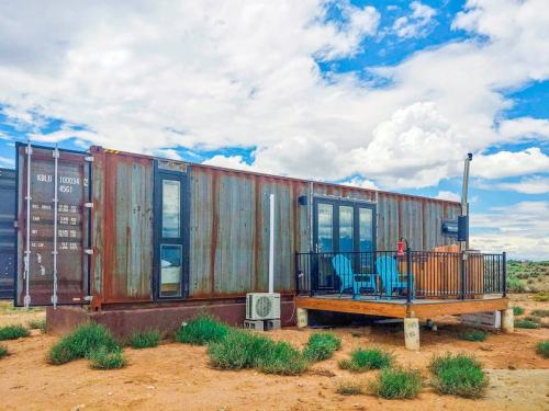 A rustic shipping container home with a wooden deck and blue chairs, set against a cloudy sky in a desert landscape.