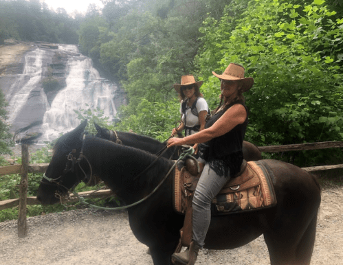 Two women in cowboy hats ride horses near a waterfall, surrounded by lush greenery.