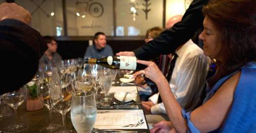 A woman points at a wine bottle during a dinner gathering with several people seated at a table.