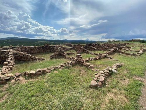 Ancient stone ruins on grassy terrain under a cloudy sky, with distant hills visible in the background.