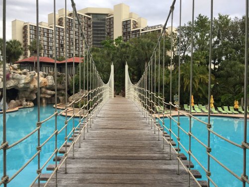 A wooden bridge spans a blue pool, surrounded by tropical plants and a hotel in the background.