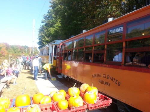 A vintage train at a pumpkin festival, with passengers boarding and pumpkins displayed in crates nearby.