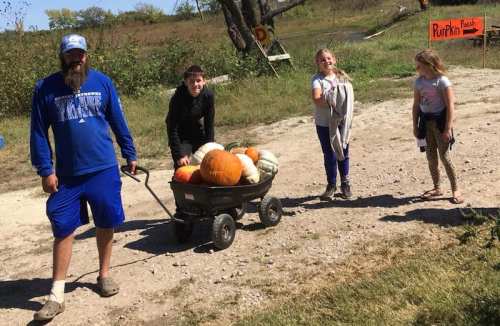 A man pulls a wagon filled with pumpkins, accompanied by three children on a sunny day at a pumpkin patch.