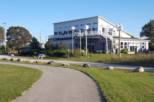 A modern building with large windows and a deck, surrounded by greenery and a winding path.