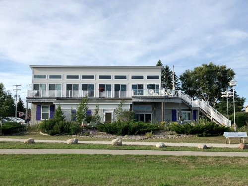 A modern two-story building with large windows, a balcony, and surrounding greenery, set against a blue sky.