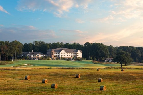 A scenic view of a golf course with hay bales in the foreground and a clubhouse in the background under a blue sky.
