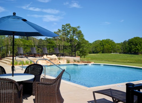 A serene outdoor pool area with lounge chairs, an umbrella, and lush greenery in the background under a clear blue sky.