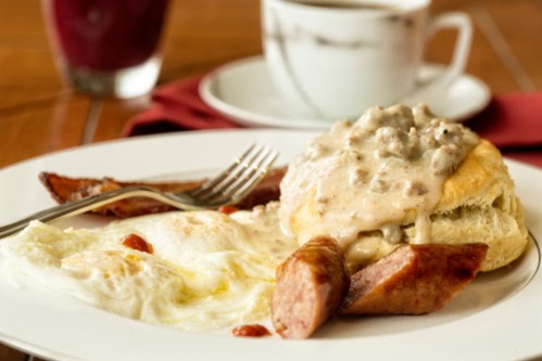 A plate of breakfast featuring eggs, sausage, a biscuit with gravy, and a cup of coffee in the background.