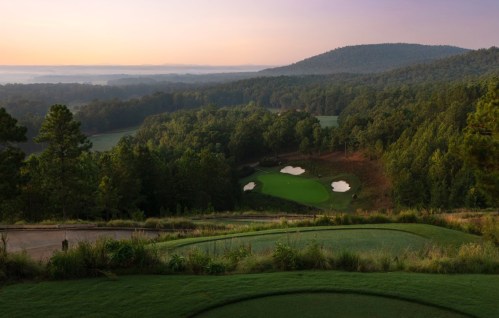 A scenic view of a golf course surrounded by lush trees and rolling hills at sunrise.
