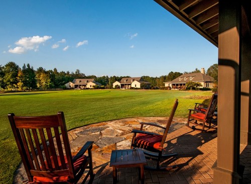 A serene view of a green lawn with rocking chairs on a porch, surrounded by trees and houses under a clear blue sky.
