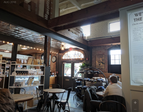 A cozy café interior with wooden beams, tables, and a counter, featuring warm lighting and a welcoming entrance.