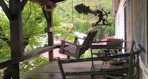 A rustic porch with a swing and a metal decoration, surrounded by greenery and a glimpse of buildings in the background.